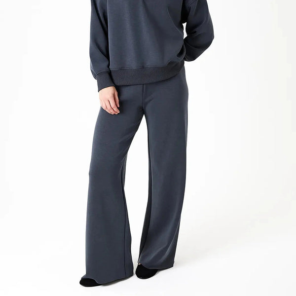 A woman modeling a dark slate gray lounge set consisting of a V-neck sweatshirt and matching wide-leg pants, photographed against a clean white studio background.